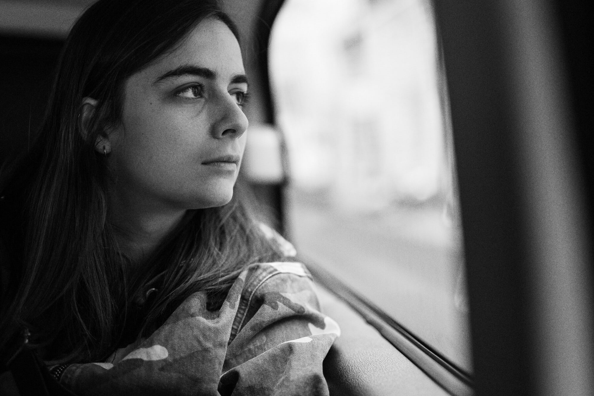 Black and white portrait of a young woman looking out the window with a serious expression, wearing a camouflage jacket.