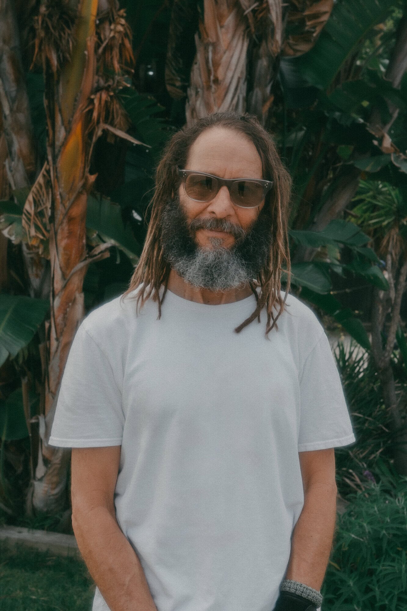 Portrait of a man with dreadlocks, sunglasses, and a beard, wearing a white t-shirt, standing outdoors with palm trees in the background.