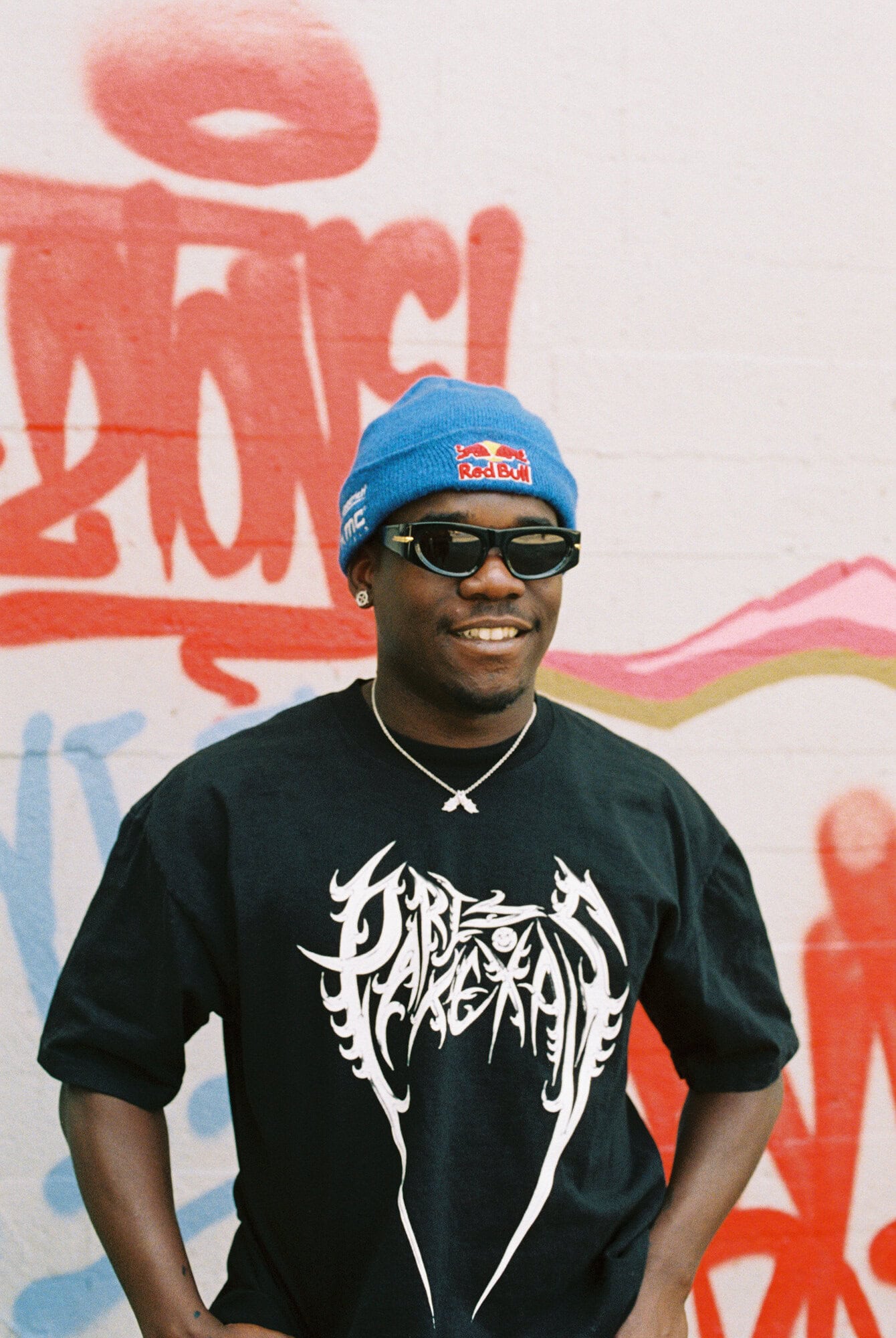 A smiling, dark-skinned man with a blue beanie, sunglasses, and black tee stands in front of graffiti art, his hands in his pockets.