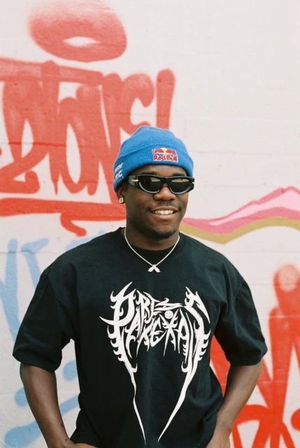 A smiling, dark-skinned man with a blue beanie, sunglasses, and black tee stands in front of graffiti art, his hands in his pockets.