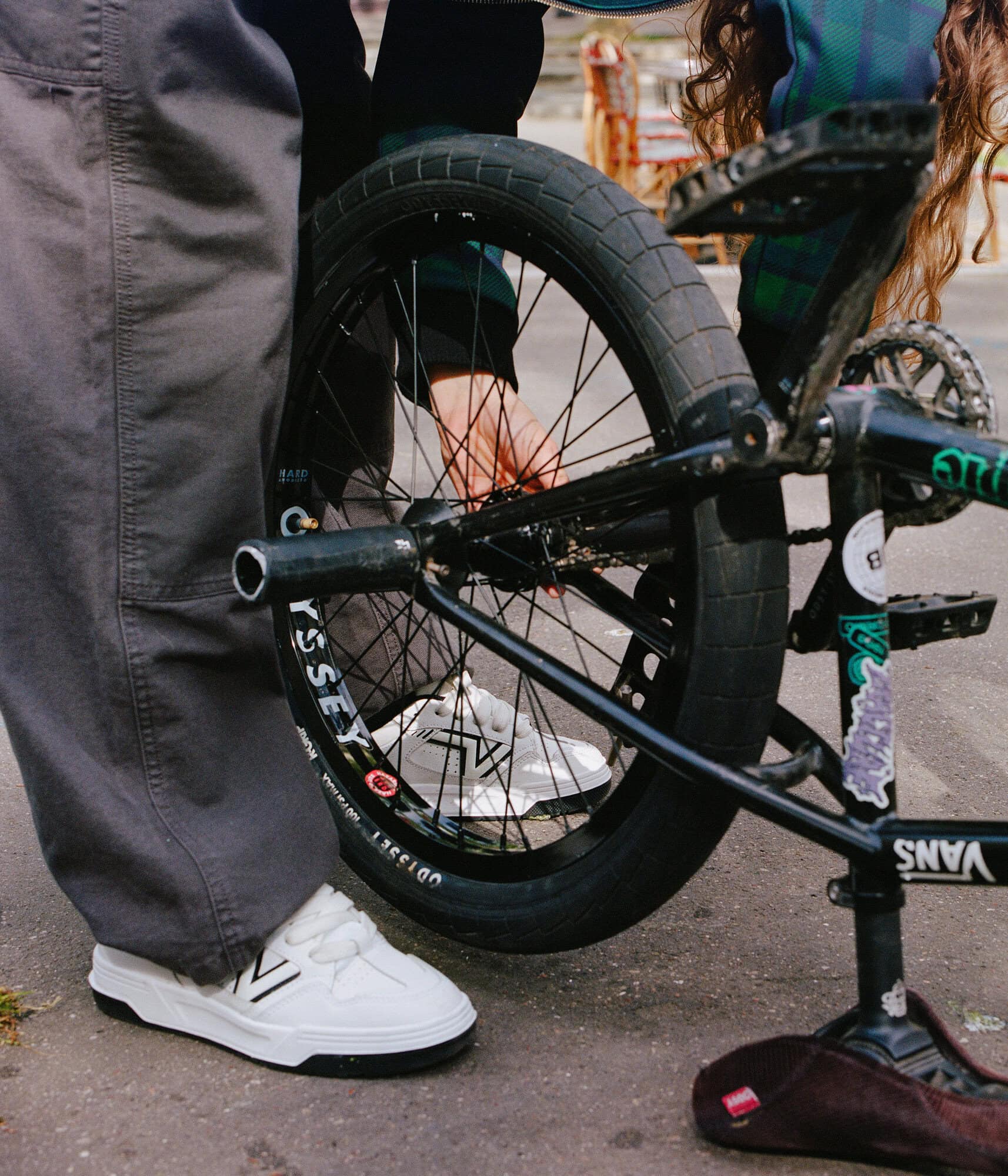 Close-up of a BMX bike's rear wheel with white Vans sneakers, gray trousers, and a green plaid jacket in an outdoor setting.