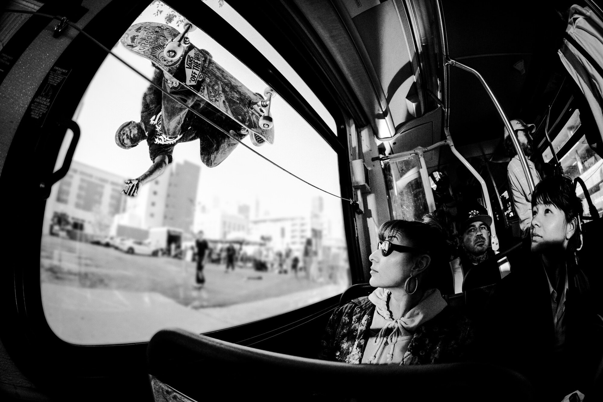 Black and white fisheye view inside a bus with a skateboarder outside the window performing a trick with urban background and bus passengers.