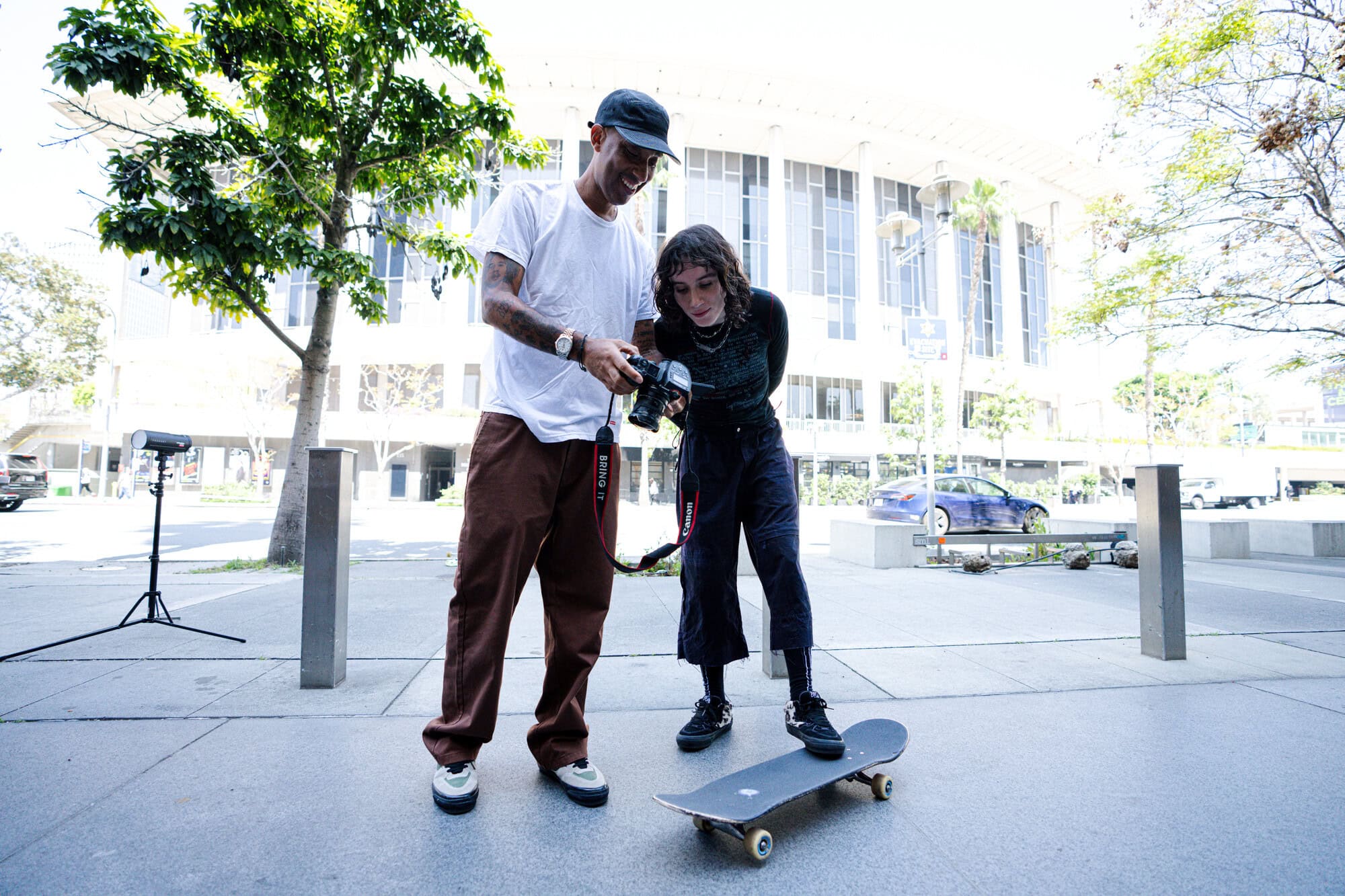 Outdoor shot of two people looking at a camera screen, one standing on a skateboard, with a building and tree in the background.