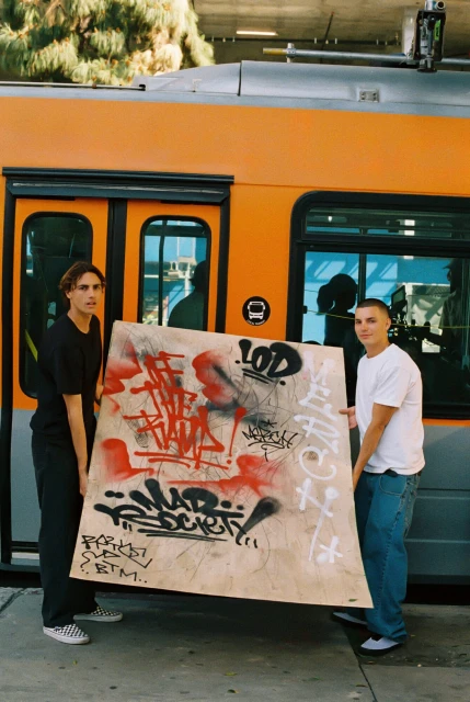 Two men hold a graffiti-covered board in front of an orange train car. The art display adds an urban element to the transportation scene.
