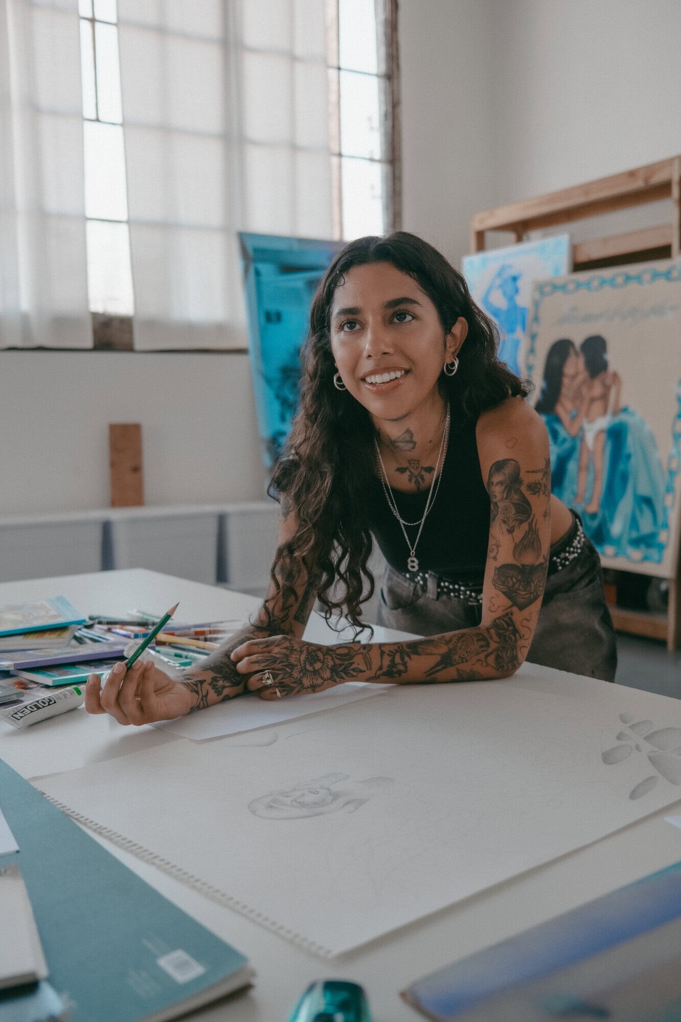 Adult female artist with tattoos sketching on a large paper in an artist studio, surrounded by art supplies and finished paintings.