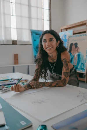 Adult female artist with tattoos sketching on a large paper in an artist studio, surrounded by art supplies and finished paintings.