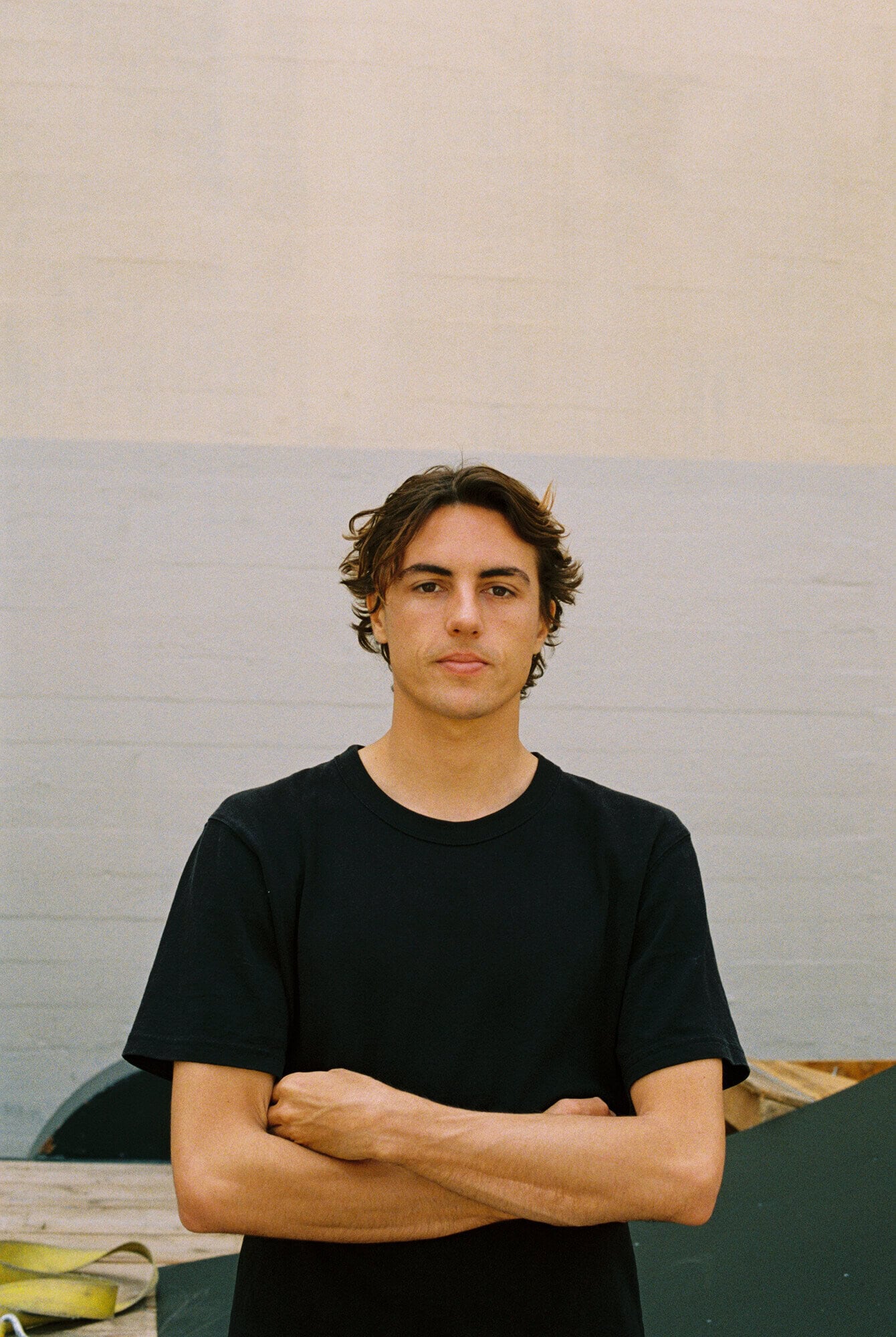 Waist-up portrait of a young adult man with dark hair posing indoors with arms crossed, wearing a black t-shirt against a neutral background.