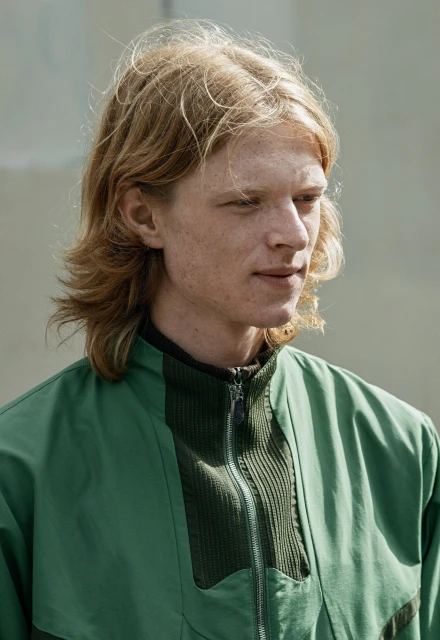 Close-up portrait of a young adult male with blonde hair wearing a green jacket, looking off to the side in an outdoor setting.