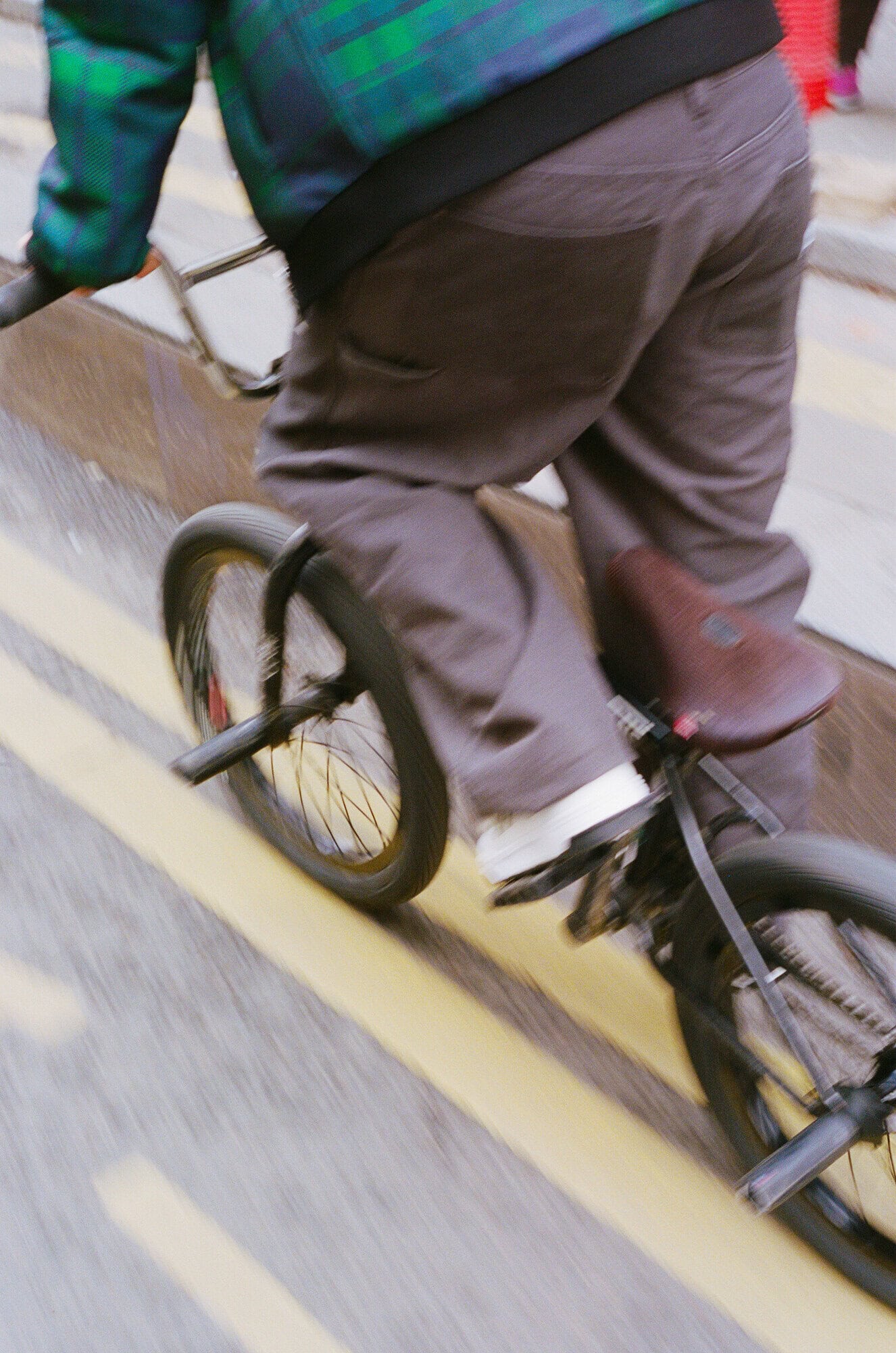 Person rides a black BMX bicycle with a brown seat on city street with yellow painted lines, motion blur, casual clothing.