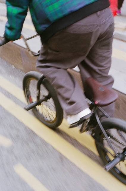 Person rides a black BMX bicycle with a brown seat on city street with yellow painted lines, motion blur, casual clothing.