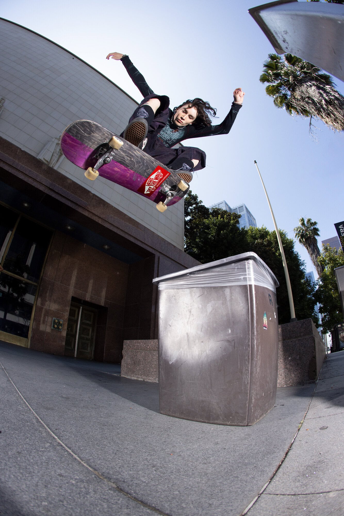 A woman performs a skateboarding trick in an urban setting. She is jumping her skateboard over a public trash receptacle.
