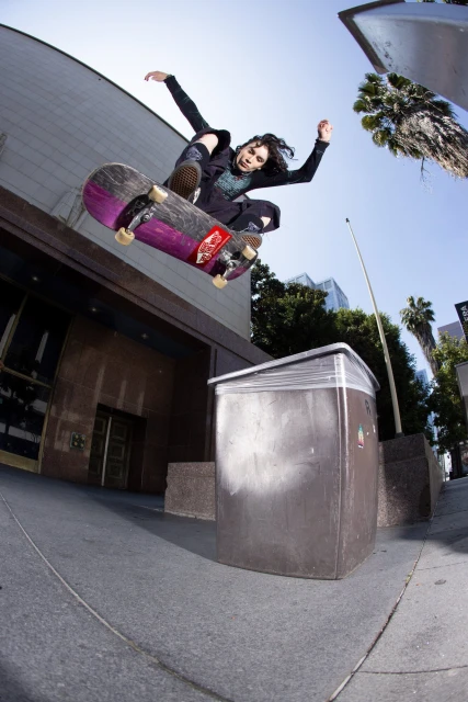 A woman performs a skateboarding trick in an urban setting. She is jumping her skateboard over a public trash receptacle.