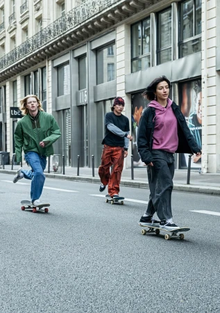 Three people skateboard down a city street lined with buildings. The focus is on the person in the front wearing a pink hoodie.