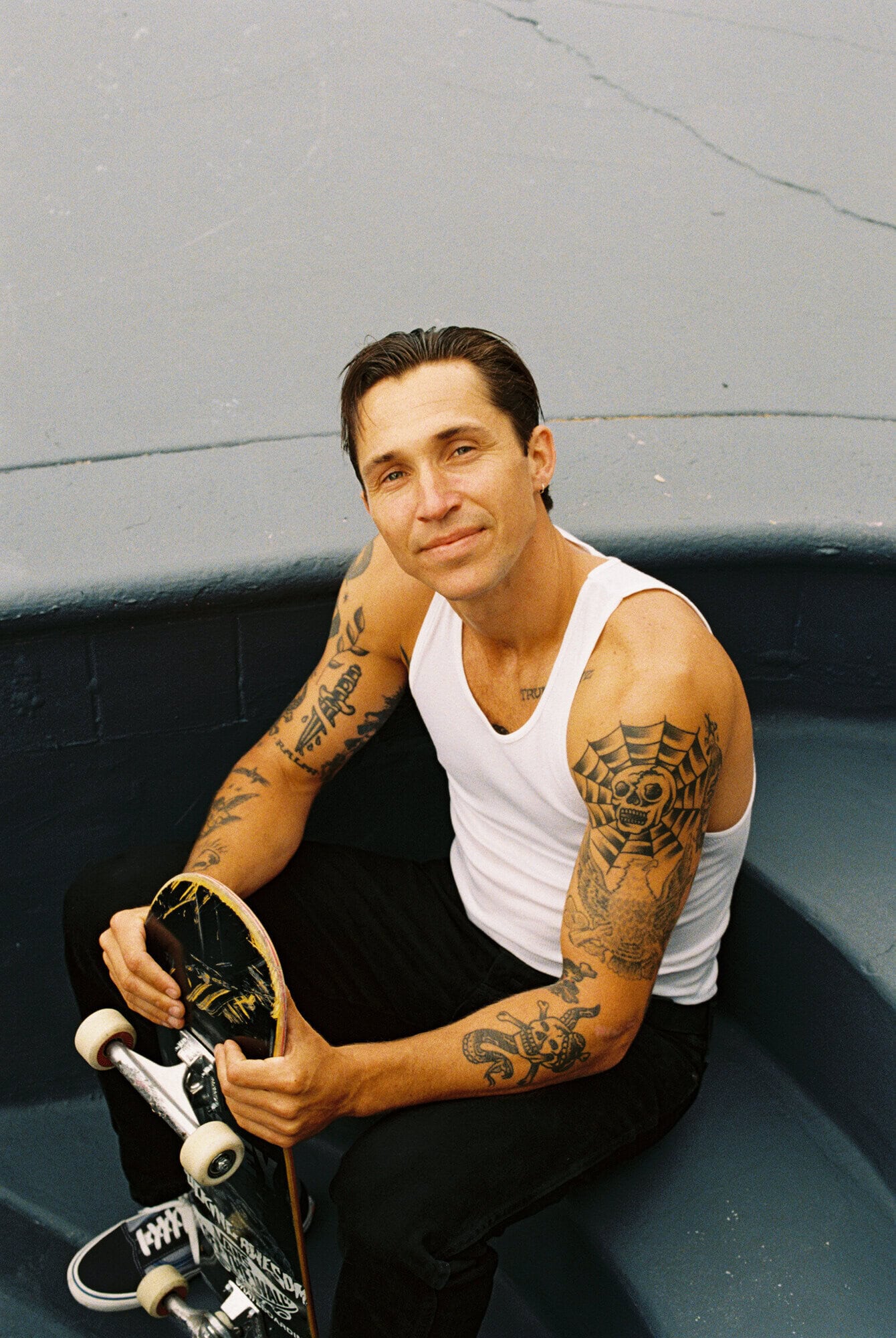 Man with arm tattoos sits holding skateboard wearing a white tank top and black pants in an outdoor skatepark setting.