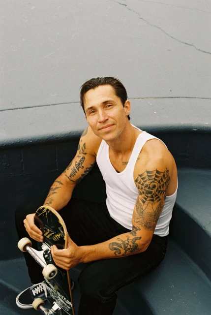 Man with arm tattoos sits holding skateboard wearing a white tank top and black pants in an outdoor skatepark setting.