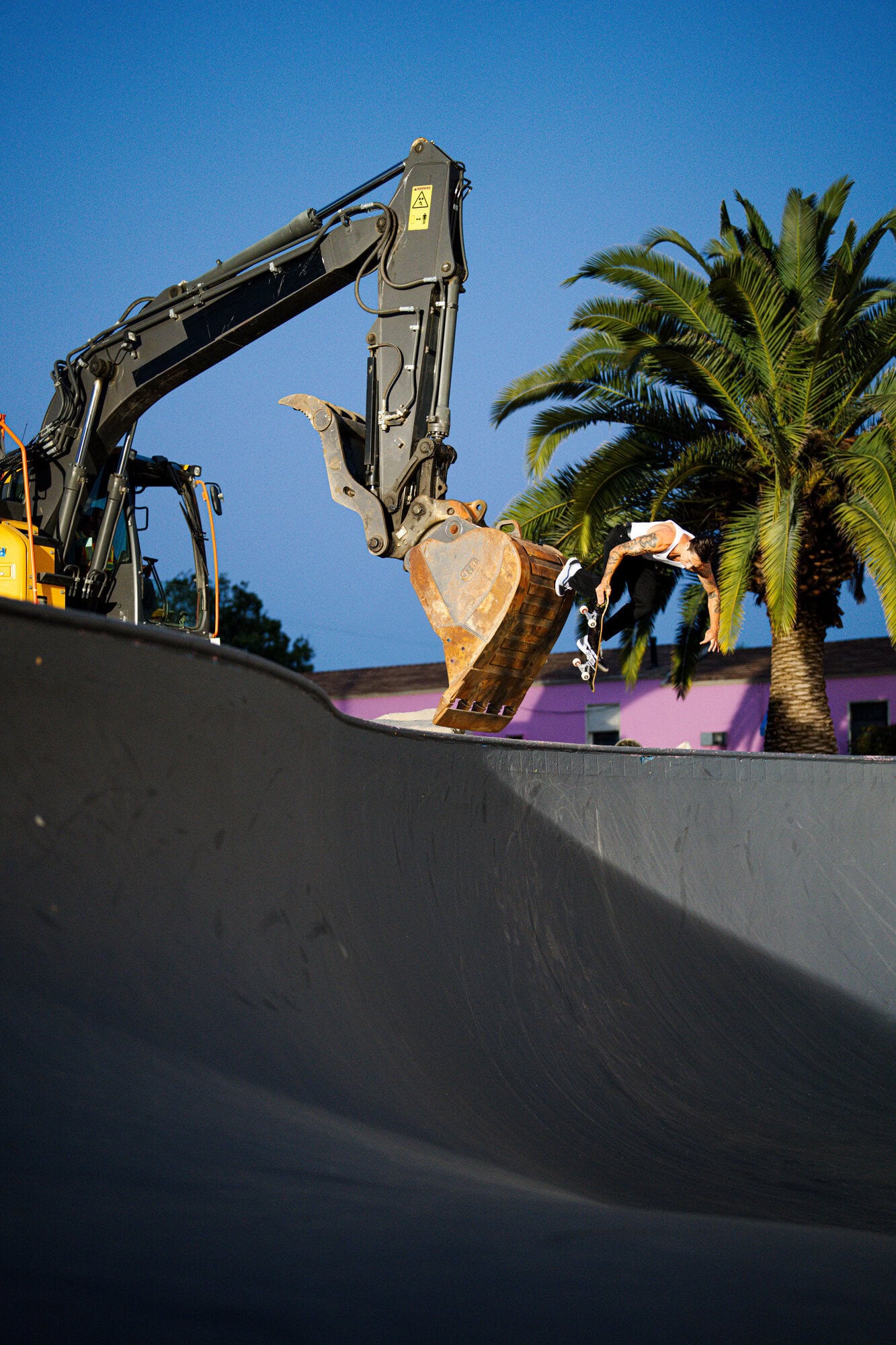 A skateboarder does a trick in a skatepark, with a large excavator arm in the background and a palm tree on a sunny day.