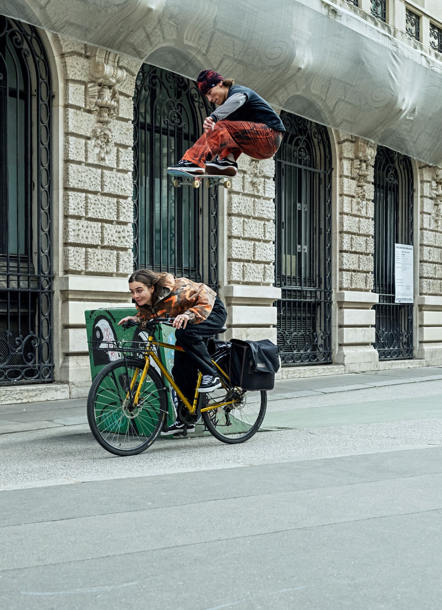 A skateboarder jumps mid-air above a cyclist on a city street in front of a stone building with arched windows and ornamental details.
