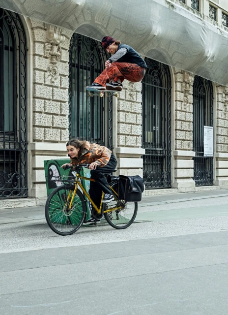 A skateboarder jumps mid-air above a cyclist on a city street in front of a stone building with arched windows and ornamental details.