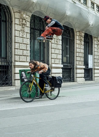 A skateboarder jumps mid-air above a cyclist on a city street in front of a stone building with arched windows and ornamental details.