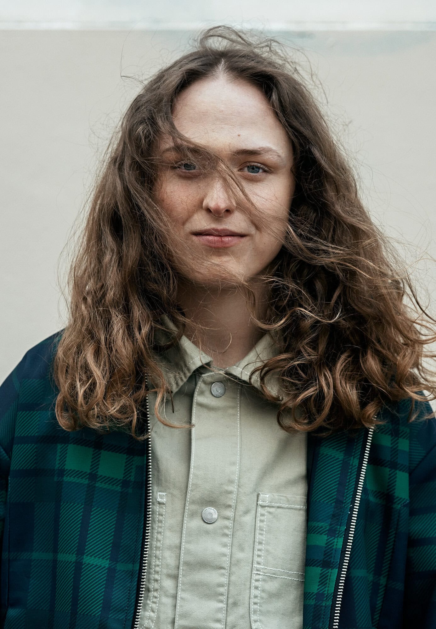 Portrait of a young adult woman with curly hair, wearing a plaid jacket and collared shirt, standing against a neutral background outdoors.