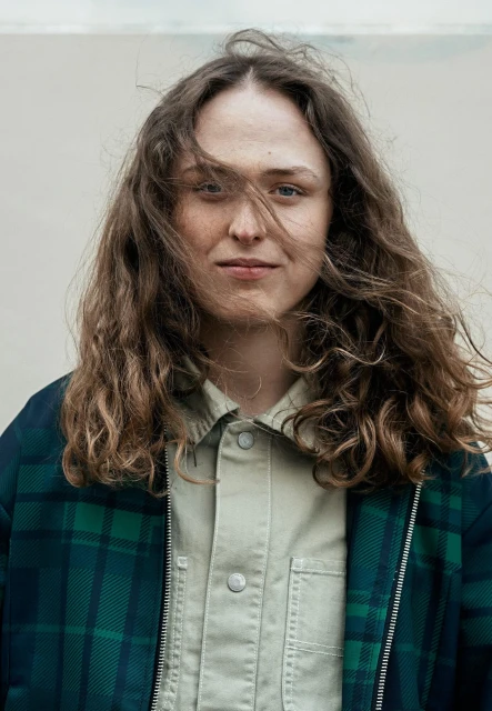 Portrait of a young adult woman with curly hair, wearing a plaid jacket and collared shirt, standing against a neutral background outdoors.