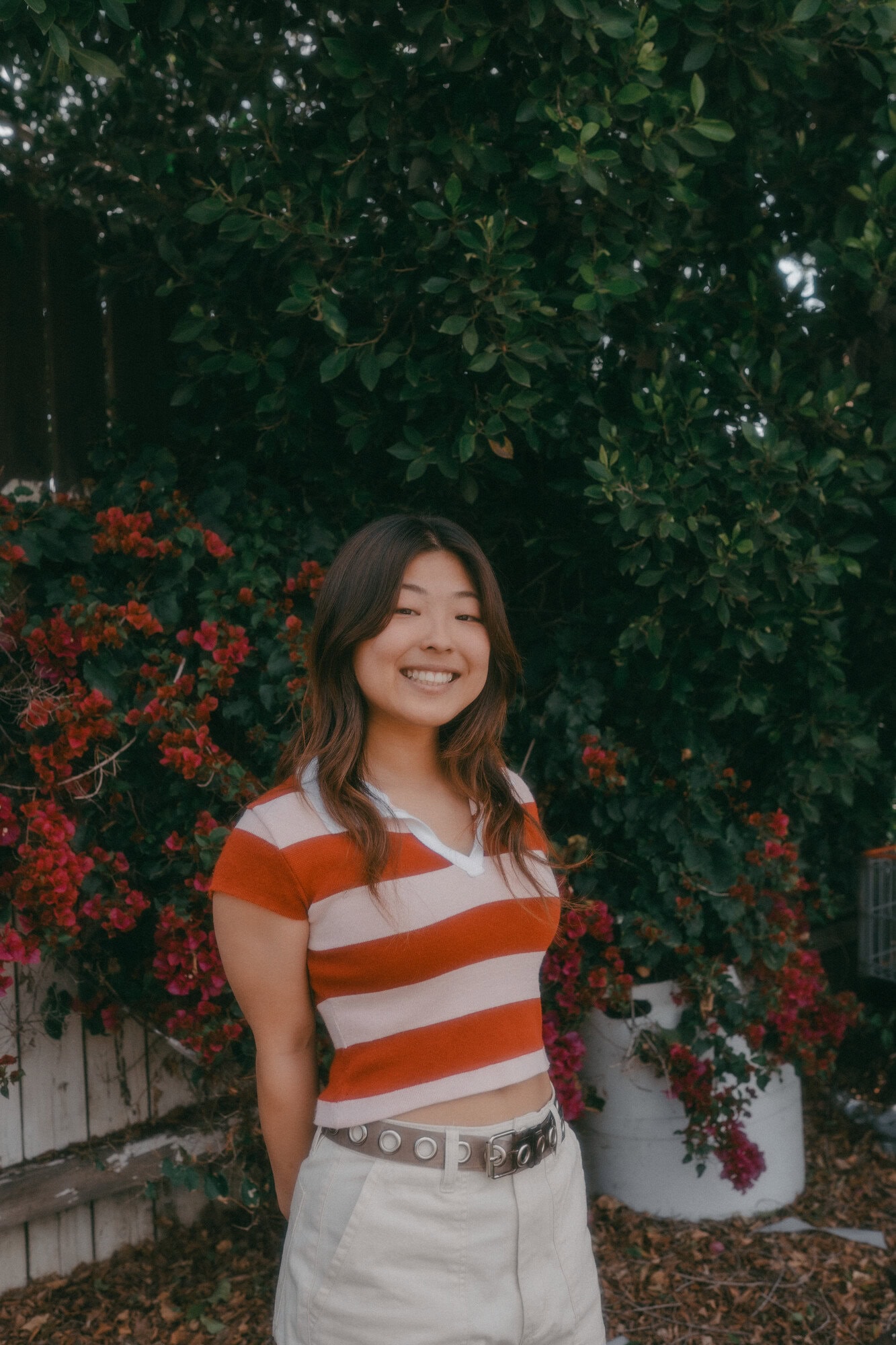 Young woman in striped orange and white crop top smiles in front of a lush green hedge with red flowers. Outdoor setting.