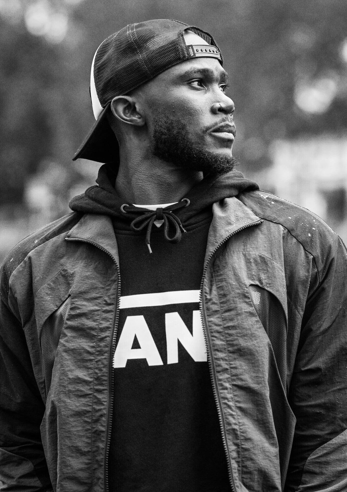 Black and white portrait of a Black man wearing a baseball cap, hoodie and jacket looking off to the side with a blurred background.