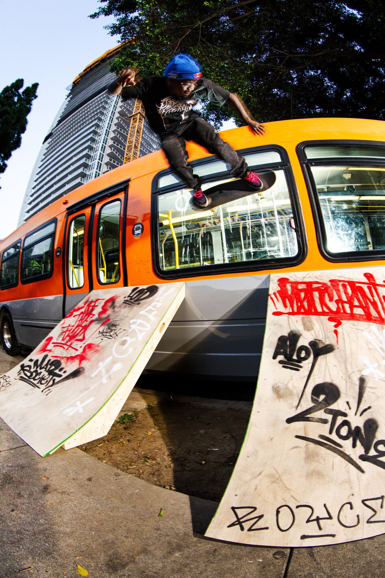 Skateboarder performs a trick on a ramp next to an orange bus in an urban setting, with a skyscraper visible in the background.