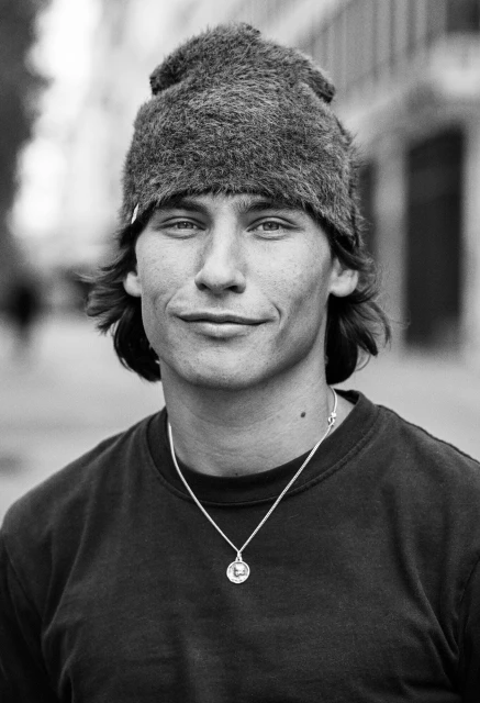 Black and white portrait of a young adult male with a slight smile, wearing a fur hat and necklace, standing outdoors in a city.