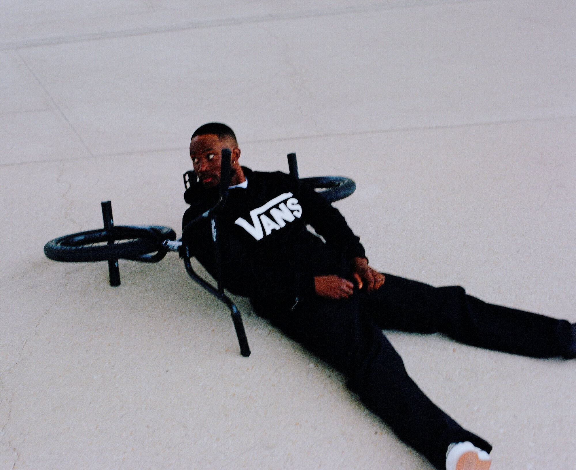 A BMX bike lies on the ground next to an African American man wearing a black Vans sweatshirt and black pants. Lying on his side, he looks at the camera.