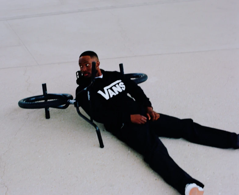 A BMX bike lies on the ground next to an African American man wearing a black Vans sweatshirt and black pants. Lying on his side, he looks at the camera.