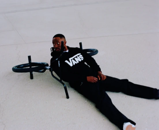 A BMX bike lies on the ground next to an African American man wearing a black Vans sweatshirt and black pants. Lying on his side, he looks at the camera.
