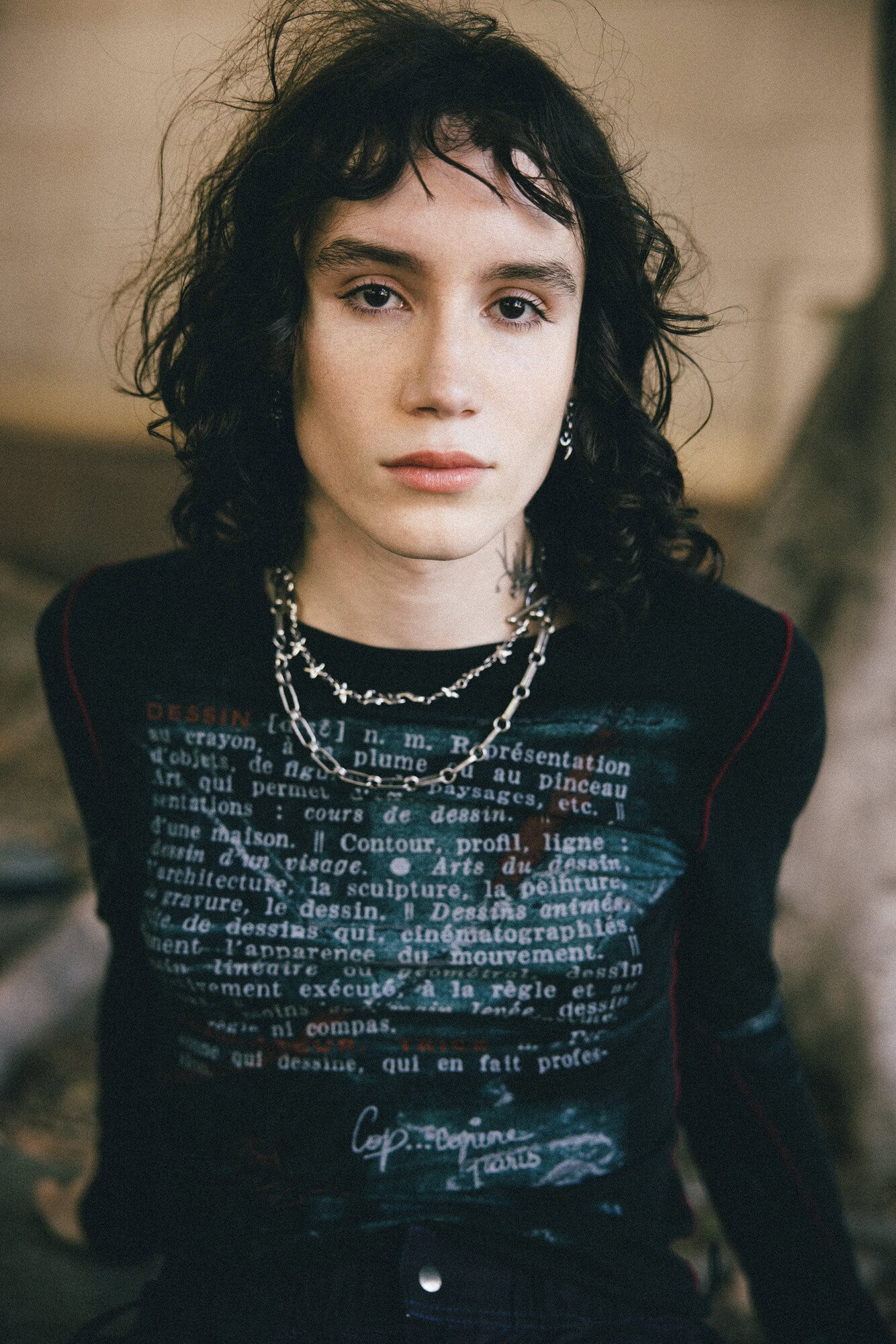 Person wearing a black graphic long-sleeve shirt with silver necklaces, posing indoors with a neutral expression and dark curly hair.