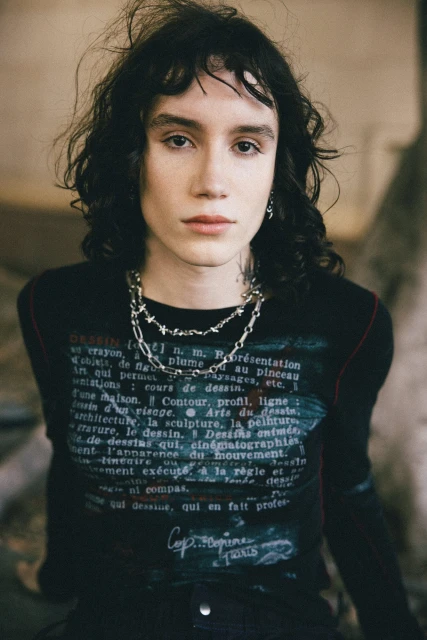 Person wearing a black graphic long-sleeve shirt with silver necklaces, posing indoors with a neutral expression and dark curly hair.
