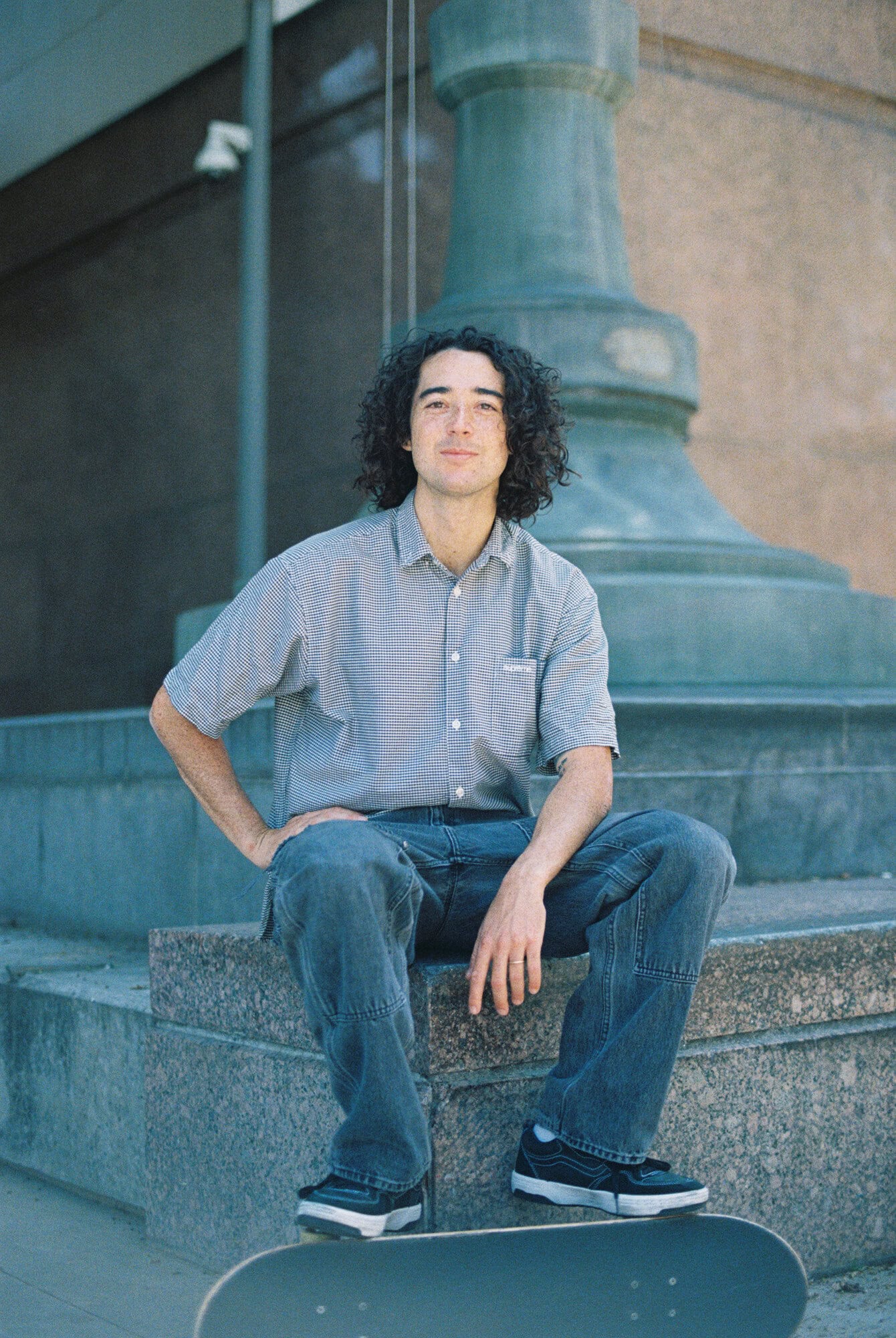 Young adult in casual attire sitting on stone steps near a building with a skateboard, captured in medium shot in outdoor setting.