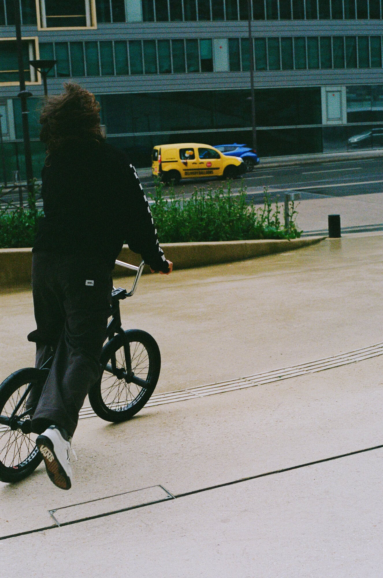 Person rides a BMX bike on a concrete plaza near an office building on a cloudy day with a yellow van driving on the road.