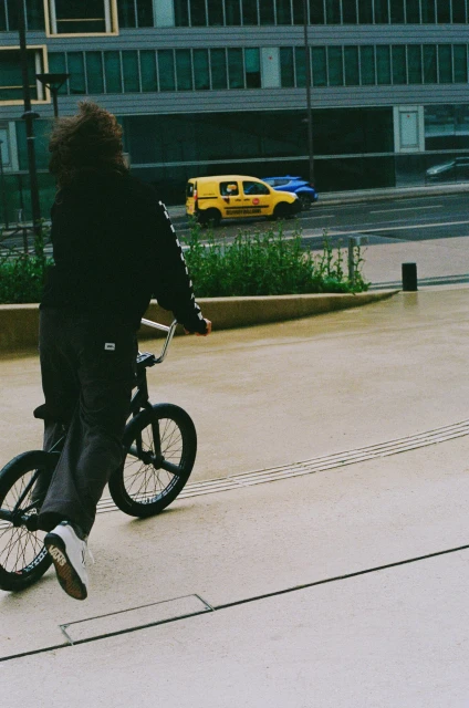 Person rides a BMX bike on a concrete plaza near an office building on a cloudy day with a yellow van driving on the road.