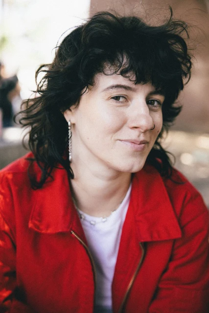Close-up portrait of an adult woman with dark curly hair wearing a red jacket and silver earrings, with an outdoor background.