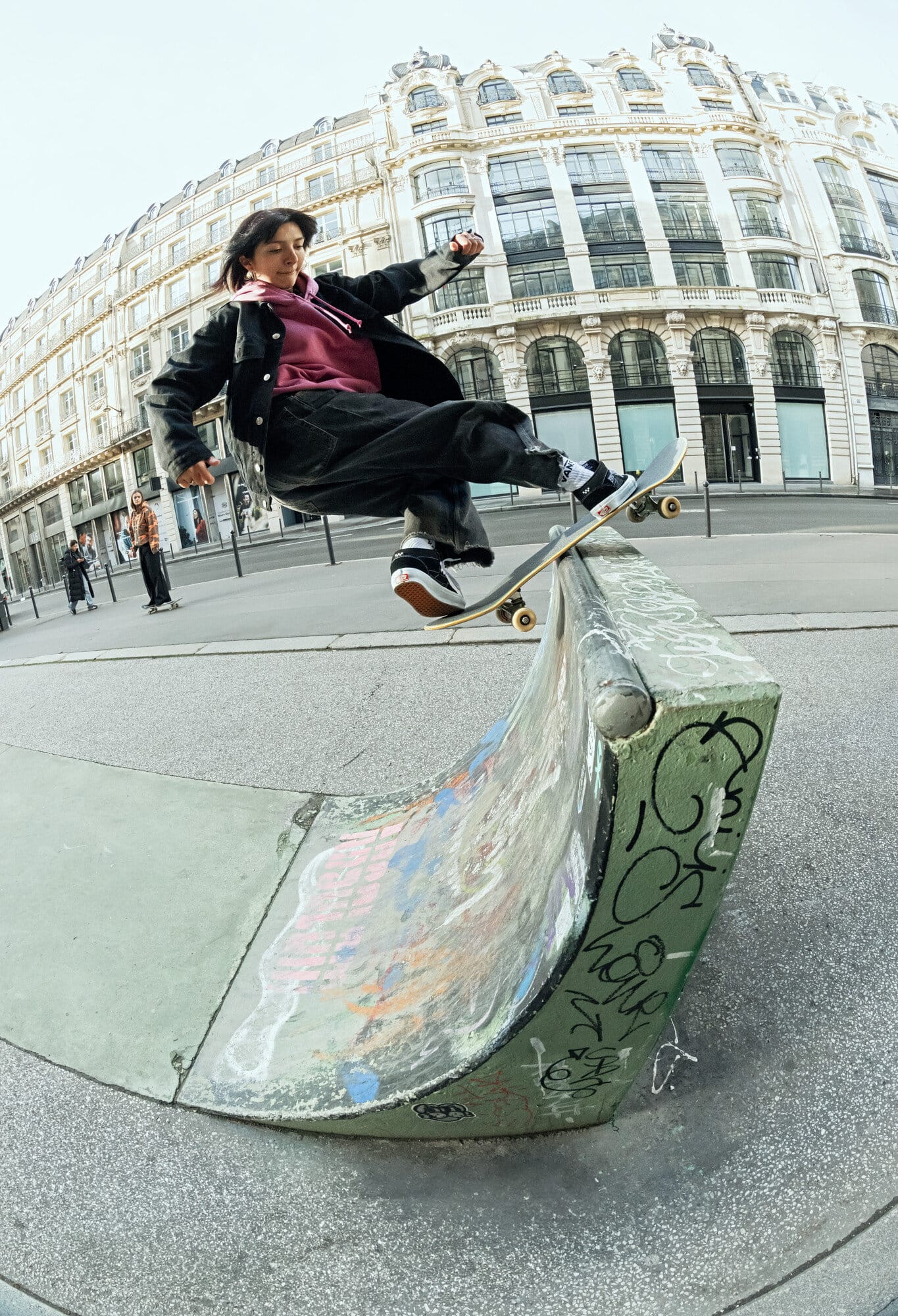 A woman on a skateboard rides a concrete ramp in an urban environment, with a blurred building in the background, captured with a fisheye lens.