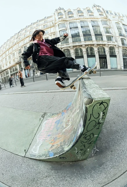A woman on a skateboard rides a concrete ramp in an urban environment, with a blurred building in the background, captured with a fisheye lens.