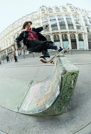 A woman on a skateboard rides a concrete ramp in an urban environment, with a blurred building in the background, captured with a fisheye lens.