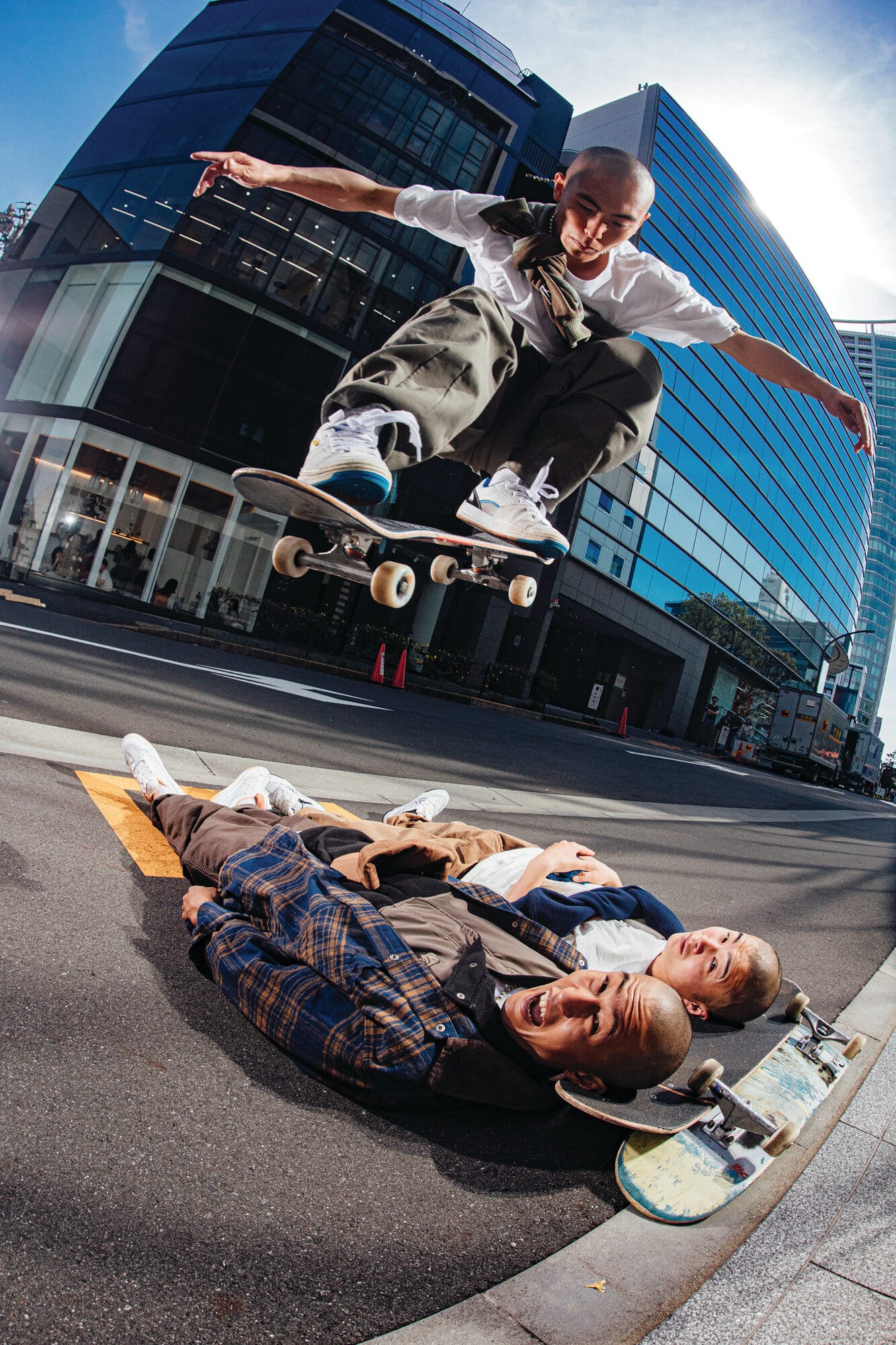 Skateboarder mid-air over two people lying on city street with modern building and clear sky in background, captured with wide-angle lens.