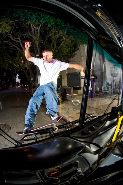Skateboarder performs a trick inside a bus with an outdoor urban setting at night, showcasing a dynamic action shot with city lights.