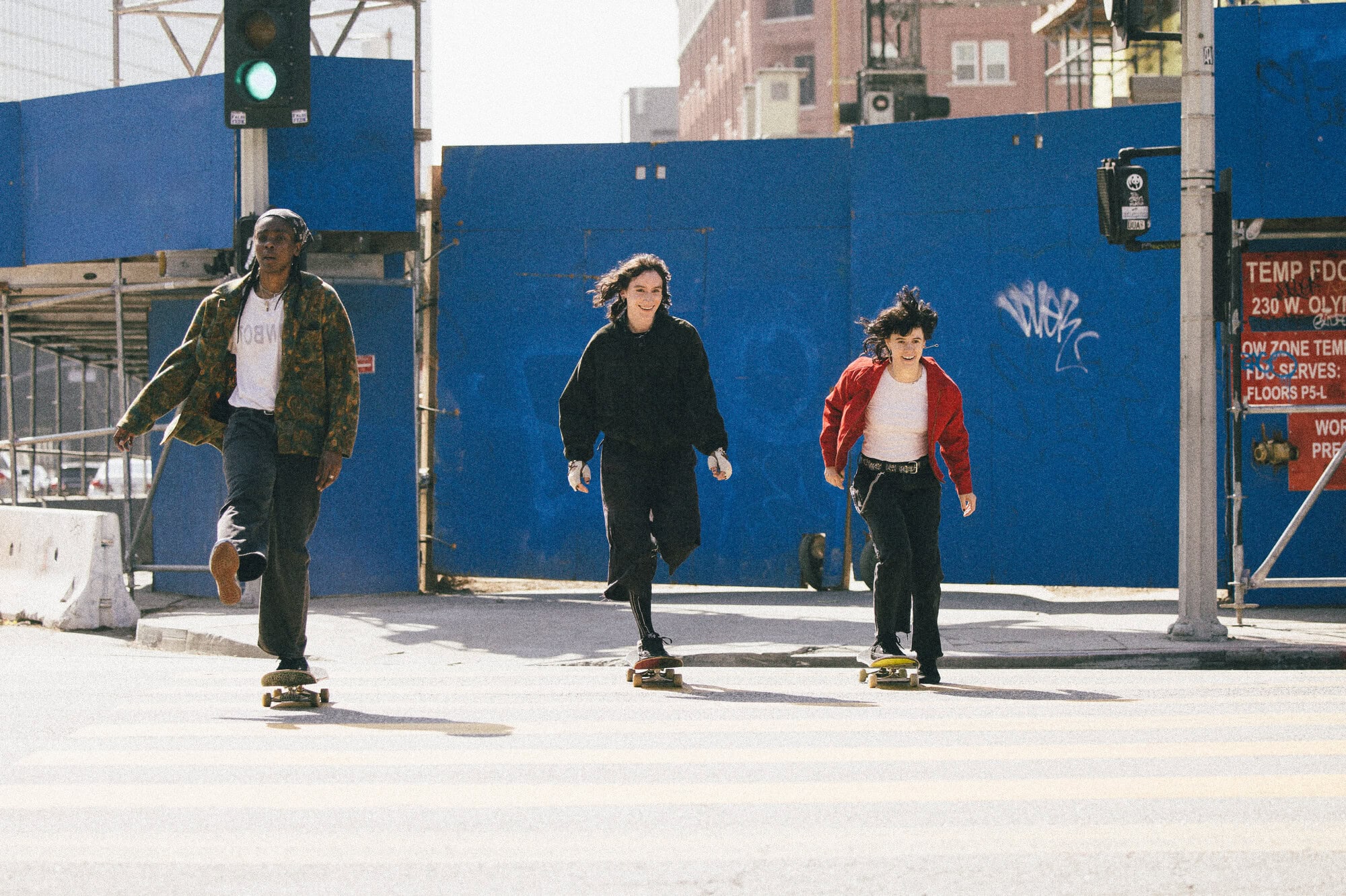 Three young women skateboarding on a crosswalk in an urban setting. Each rides a skateboard wearing casual clothing. Bright daylight. 