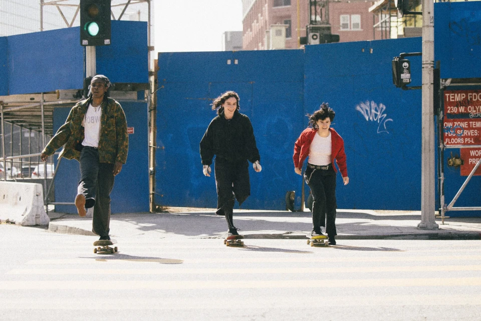 Three young women skateboarding on a crosswalk in an urban setting. Each rides a skateboard wearing casual clothing. Bright daylight. 