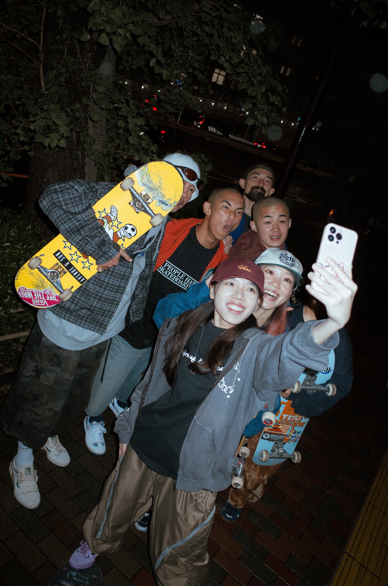 Group of six Asian adults and youths taking a selfie on an urban sidewalk at night, holding skateboards, wearing streetwear.