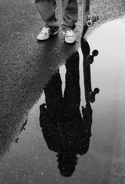 Black and white shot of a skateboarder reflected in a puddle, showing feet, skateboard, jeans, and pavement.