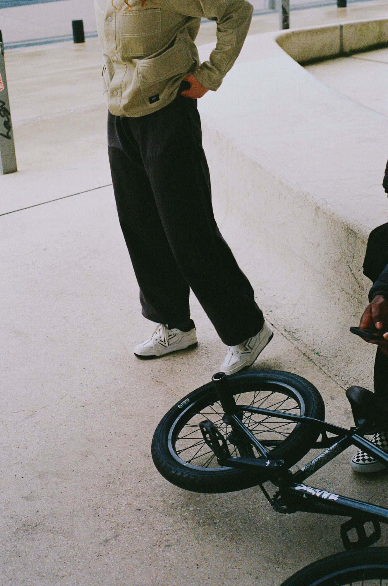 Person standing near a BMX bike in a skate park with a concrete surface, wearing casual clothes and white sneakers.