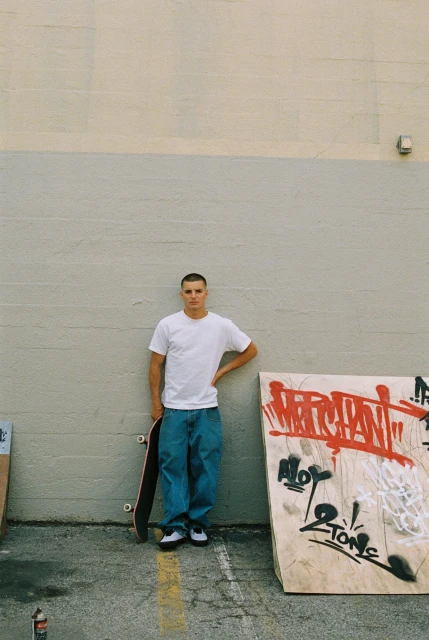 A young adult man in a white t-shirt and jeans poses with a skateboard outdoors against a concrete wall with graffiti in daylight.