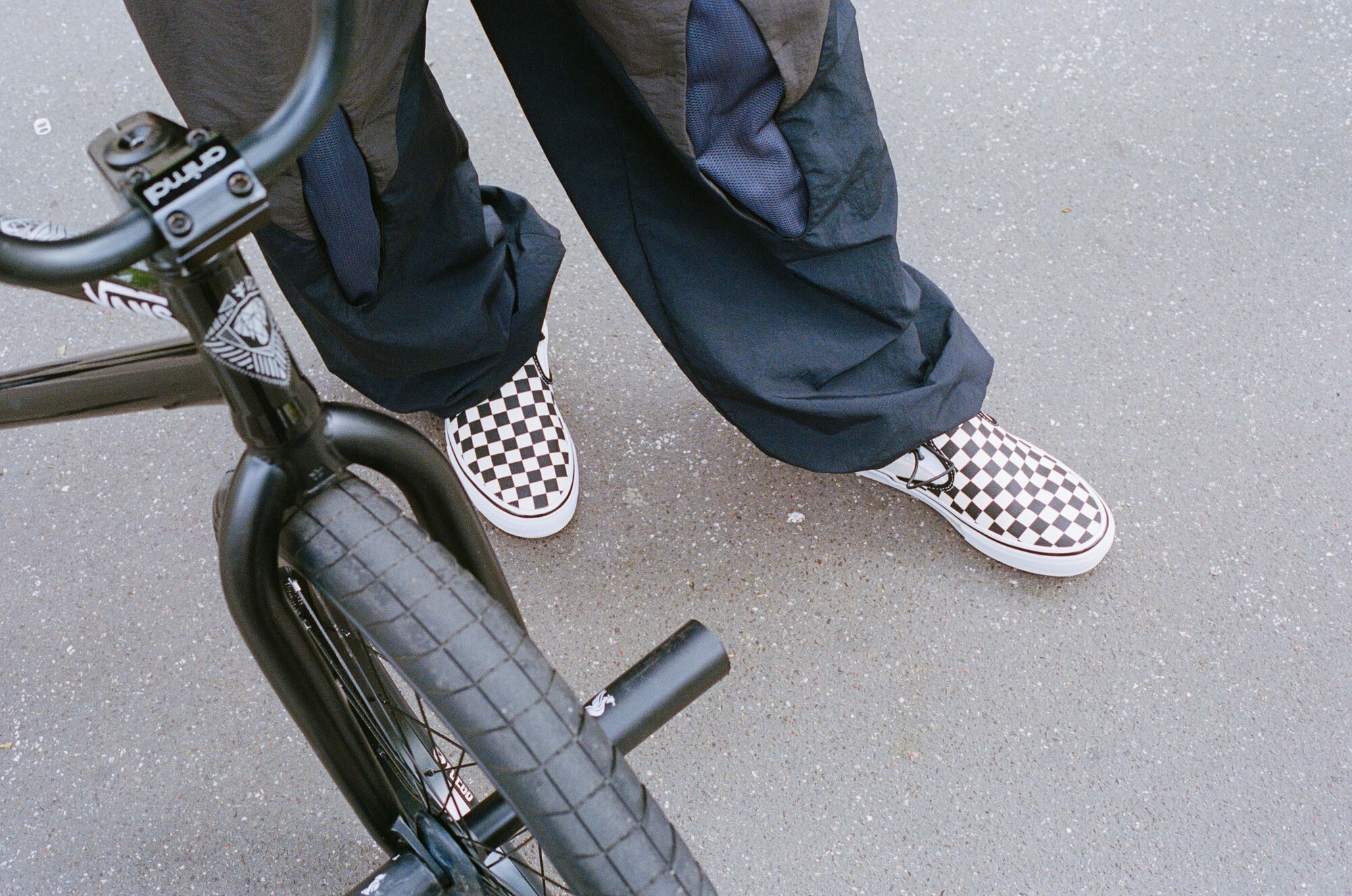 Person wearing checkered Vans with a black BMX bike on grey pavement, showcasing urban style and action sports lifestyle.
